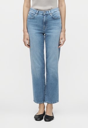 Személy világoskék egyenes szárú farmernadrágot visel gombbal és cipzárral, fekete lapos cipővel párosítva, fehér háttér előtt áll.