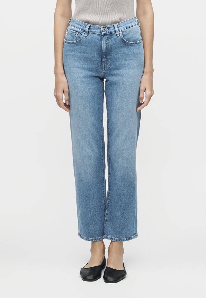 Άνθρωπος που φορεί ανοιχτό μπλε τζιν με ίσια γραμμή, κουμπί και φερμουάρ, συνδυασμένο με μαύρα μπαλαρίνες, στέκεται μπροστά σε λευκό φόντο.