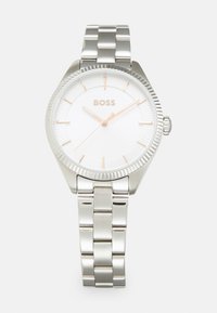 Sølvklokke med en teksturert hvit skive og rose gull detaljer. Har en armbånd av rustfritt stål og en ribbet bezel.