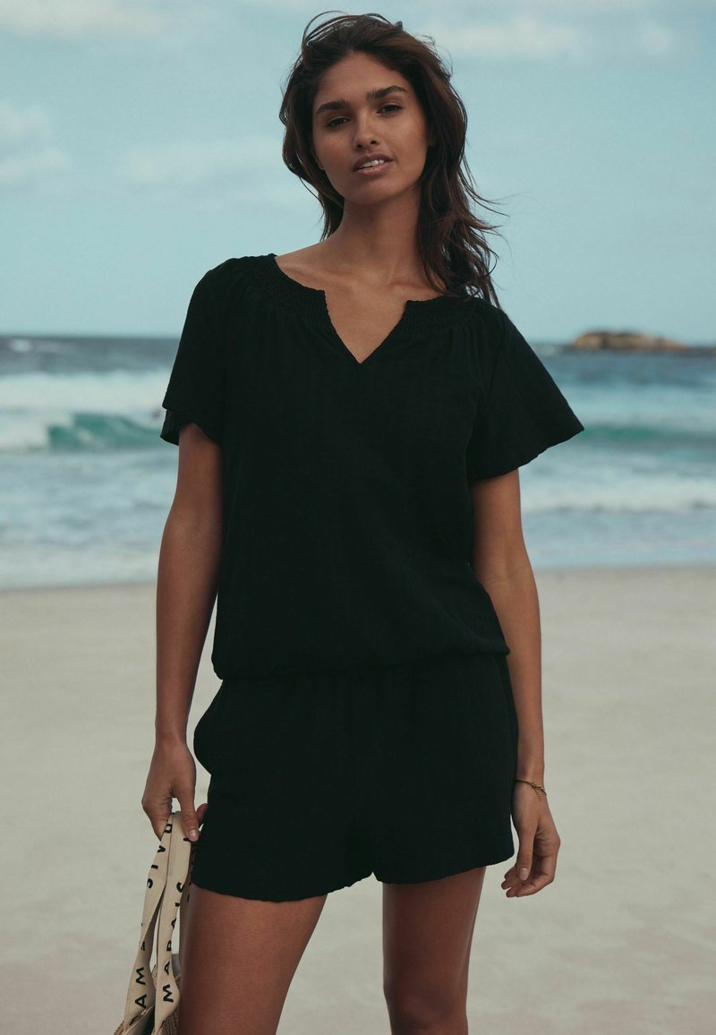 Schwarzes kurzärmeliges Oberteil mit V-Ausschnitt und Shorts mit elastischem Bund. Glatte Textur; Model hält eine beige Tasche mit schwarzer Schrift. Strand-Hintergrund.