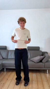 Cream cotton t-shirt with a small bird graphic, paired with black trousers. Short sleeves, fitted shape, worn with dark shoes. Wooden floor.