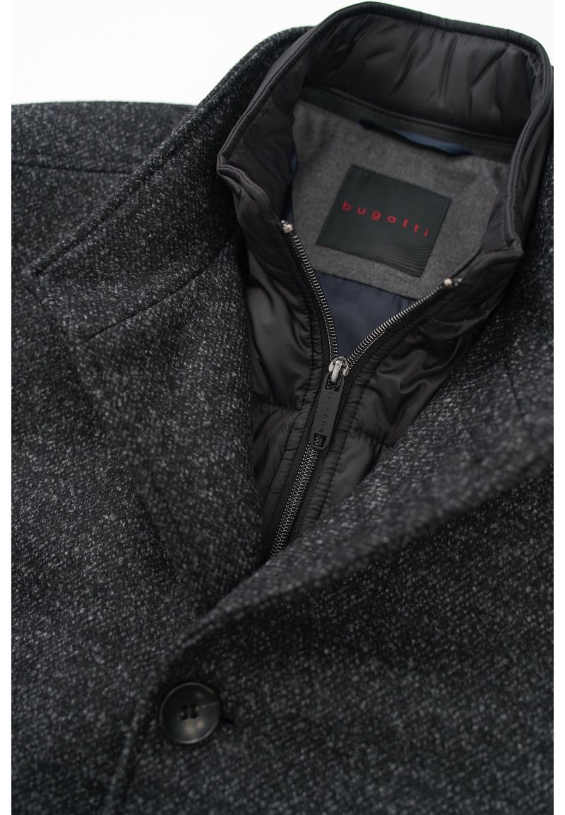 Cappotto grigio scuro in misto lana con una fodera nera trapuntata, dotato di un colletto a punta, chiusura con zip e un evidente dettaglio con bottone. Etichetta visibile.