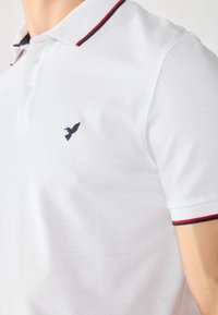 Valge polo särk tekstureeritud kangaga, millel on tumesinine tikitud linnu logo ja krae, millel on punased ja tumesinised triibud.