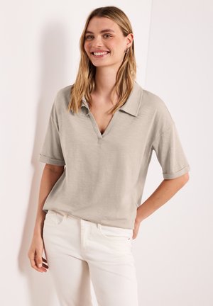 Femme souriante aux cheveux blonds mi-longs porte un haut beige à manches courtes avec col et un pantalon blanc, debout contre un mur clair.