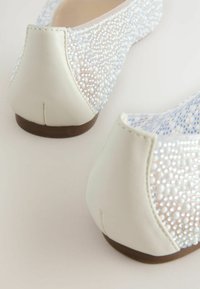 Chaussures plates blanches avec un dessus texturé en perles, mettant en avant des accents en cuir lisse et un bout arrondi. Semelle plate marron, sans quincaillerie visible.