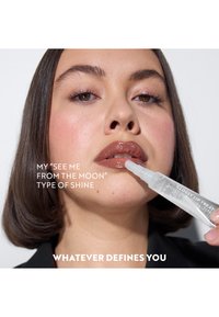 Gjennomsiktig leppglansrør med en avsmalnende applikator, med blank brune lepper. Modellen har mellomlangt hår og nøytral makeup mot en hvit bakgrunn.