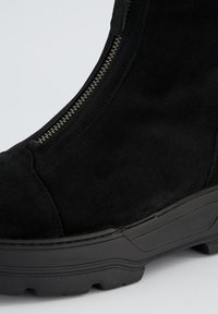 Bottine en suède noir avec fermeture éclair latérale et semelle en caoutchouc avec des détails texturés, conçue pour la durabilité et le confort.