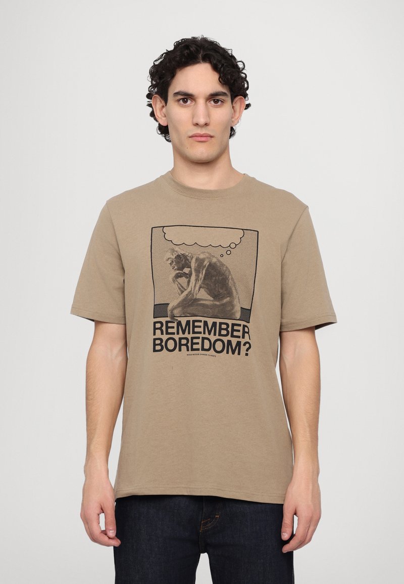 T-shirt en coton beige avec un col ras du cou, présentant un imprimé graphique d'une silhouette pensive et le texte "TE SOUVIENS-TU DE L'ENNUI ?" en noir.