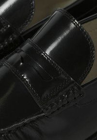 Gros plan sur des mocassins penny en cuir noir poli avec des coutures détaillées et une bride classique sur le dessus.