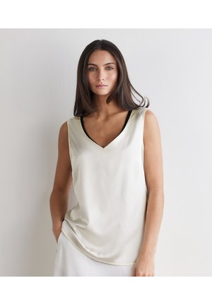 Frau mit langen dunklen Haaren, die ein ärmelloses, cremefarbenes V-Ausschnitt-Top trägt und vor einer schlichten weißen Wand steht, die Hände teilweise in den Taschen.