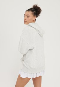 Haut de survêtement oversize gris clair avec une capuche à cordon, des manches longues et une pochette, porté sur une jupe plissée blanche. Texture lisse.