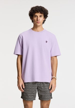 Light purple cotton t-shirt with short sleeves and a round neck, featuring a small dark emblem on the chest, paired with patterned black shorts.