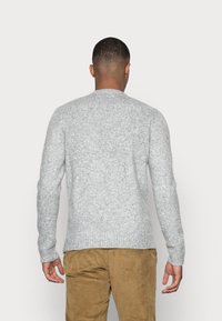 Uomo con i capelli corti, che indossa un maglione lavorato a maglia grigio chiaro e pantaloni di velluto a coste marroni, in piedi di spalle contro uno sfondo semplice.