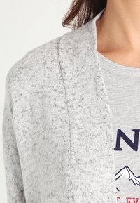 Jasnoszary kardigan z teksturowanym wzorem dzianiny i otwartym przodem. Nałożony na szary T-shirt z grafiką gór w granatowym kolorze.
