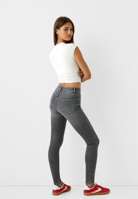 Szürke farmer skinny nadrág hátsó zsebekkel, fehér fitt toppal és piros sportcipővel párosítva. A modell karjait összekulcsolva, kissé hátrafordulva pózol.