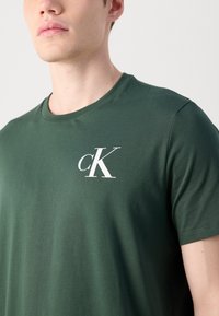 Grøn bomulds T-shirt med rund halsudskæring og korte ærmer, der har et hvidt "cK"-logo på venstre brystområde.