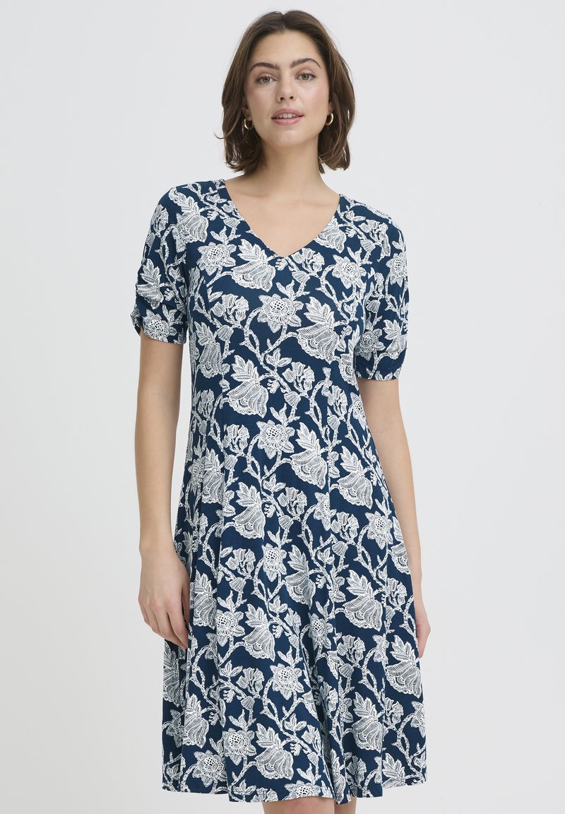 Kobieta w niebieskiej sukience do kolan z białym kwiatowym wzorem i krótkimi rękawami, stojąca na jednolitym jasnym tle.