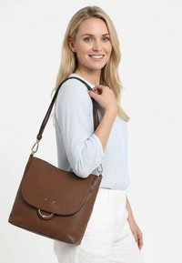 Naeratav naine, kes kannab helesinist pluusi ja valgeid pükse, kandes keskmisepruunist nahast õlakotti, millel on hõbedased rõnga detailid.