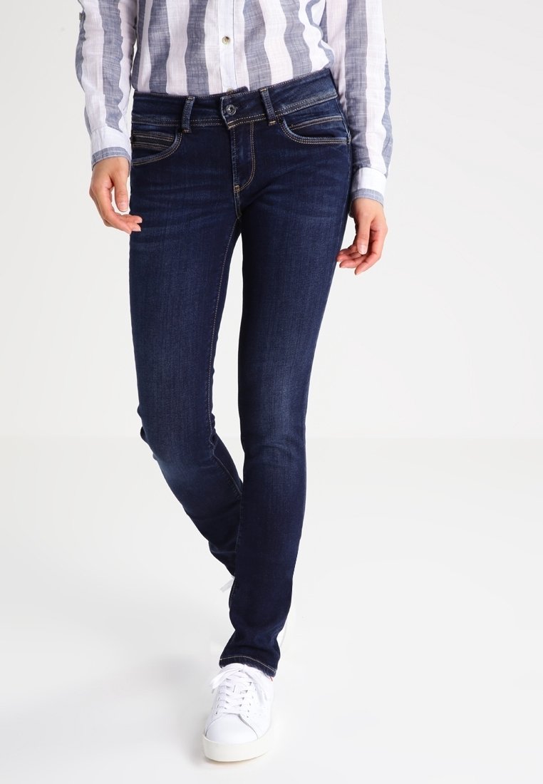 Vaqueros ajustados de color azul oscuro con un corte clásico, cinco bolsillos y costuras en contraste, combinados con zapatillas blancas y una camisa de botones a rayas.