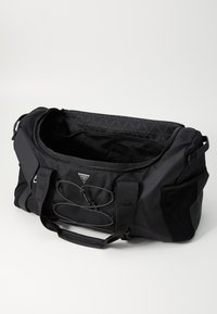 Sac de sport noir avec un compartiment principal zippé, des accents en maille et une sangle ajustable. Marquage notable et détail de couture décorative.