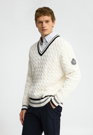 Joven con un suéter blanco de punto con cuello en V y ribete negro y una camisa de rayas con cuello, de pie con una mano en el bolsillo.
