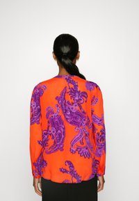 Blusa a maniche lunghe in tessuto arancione vivace con un motivo paisley viola. La vista posteriore mostra un taglio dritto con polsini con bottoni.