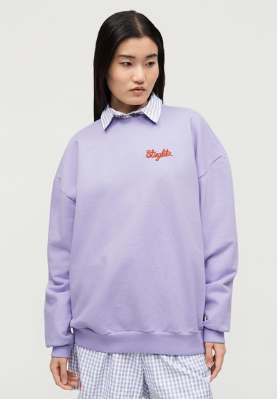 Γυναίκα με μακριά σκουρόχρωμα μαλλιά, που φοράει ένα φαρδύ λιλα φούτερ με λογότυπο "Stieglitz" και κάτω από αυτό ένα μπλε πουκάμισο με καρό μοτίβο gingham.