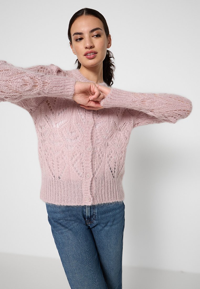 Różowy kardigan wykonany z dzianiny o fakturze z liściastym wzorem, z długimi rękawami i zapięciem na guziki, połączony z niebieskimi dżinsami.