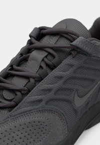 Gros plan d'une basket Nike gris foncé montrant la tige en mesh texturé, l'empeigne en daim, les lacets croisés et le logo swoosh latéral.