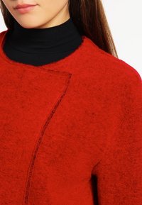 Punainen villaneule, jossa on tekstuuri, pyöreä kaula-aukko ja erottuva limittäin oleva etuosan muotoilu, päällä musta poolopaita.