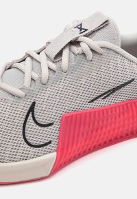 Zapatilla atlética de color gris claro con parte superior de malla, acentos negros y una suela de goma rosa que presenta un diseño texturizado. Los cordones son planos y suaves.