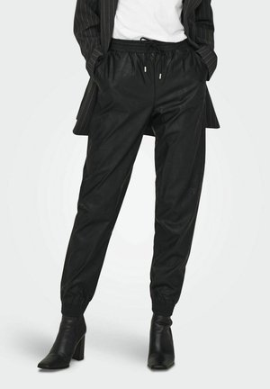 Kobieta ubrana w czarne joggery z ekoskóry z elastycznymi mankietami, czarne botki na obcasie, biały top i marynarkę w drobną prążkowaną kratę, ręce w kieszeniach.