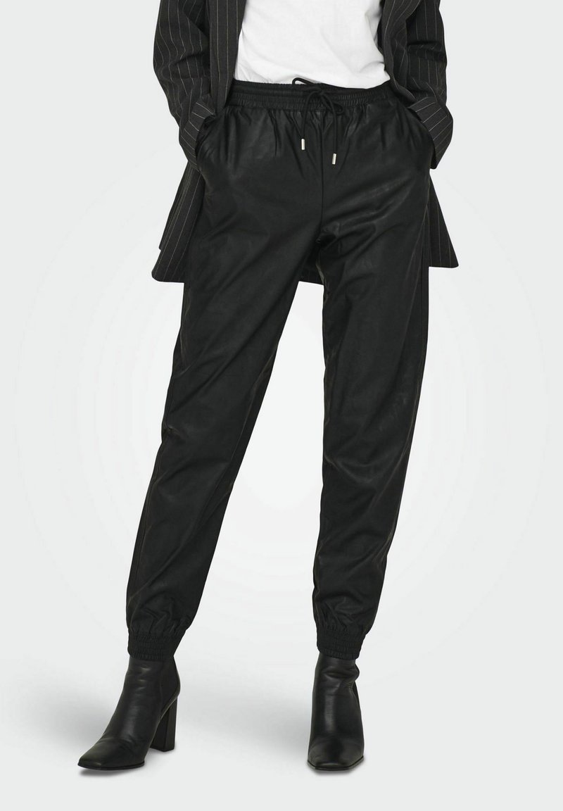 Femme portant un pantalon jogger en similicuir noir avec des ourlets élastiques, des bottines à talons noires, un haut blanc et un blazer rayé, les mains dans les poches.