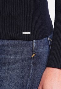 Närbild av en person som bär en marinblå ribbad tröja och blå denimjeans med en silverfärgad rektangulär metalltagg på tröjan.