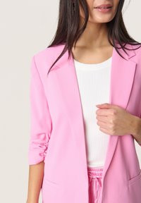 Rosa Blazer aus glattem Stoff mit hochgerollten Ärmeln und tailliertem Revers. Über einem weißen, gerippten Oberteil getragen. Dezente Nähdetails sichtbar.