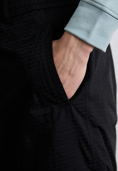 Musta püksid tekstuuritud pinna ning "Emporio Armani" logo. Reguleeritav vöökoht ja küljetasku detail on nähtavad.