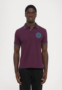 Άνδρας που φοράει ένα μωβ πόλο μπλουζάκι με μπλε περιθώριο και λογότυπο σε κυκλικό σχήμα "Versace Jeans Couture" στο στήθος, στέκεται μπροστά σε λευκό φόντο.