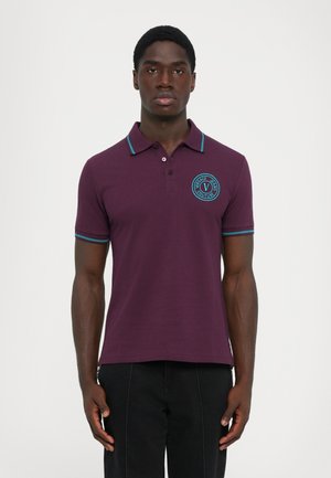 Egy férfi lila poloinget visel kék díszítéssel és a mellkasán egy kör alakú "Versace Jeans Couture" logóval, fehér háttér előtt áll.