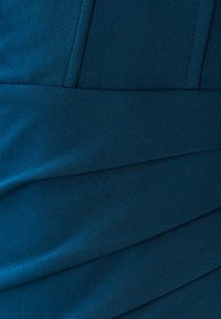 Sinihtävänvihreä kangas, jossa on sileä pinta, pystysuorat paneelit ja vinot liehutusyksityiskohdat, luoden jäsennellyn siluetin.