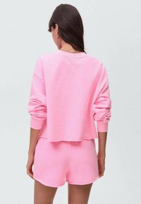 Roza skrajna dukserica z rebrastimi manšeti, v kombinaciji z ujemajočimi se kratkimi hlačami. Tkanina se zdi mehka in teksturirana, z zasnovo sproščenega kroja.