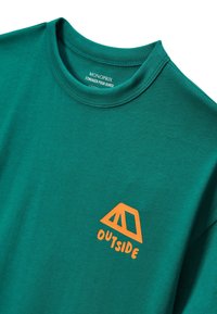 T-shirt vert avec un graphisme de tente orange et le mot "DEHORS" imprimé en dessous sur la poitrine gauche.