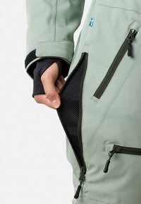 Wodoodporna kurtka w jasnozielonym materiale. Posiada zamknięcia na zamek błyskawiczny, czarną siateczkową podszewkę, regulowane mankiety oraz emblemat szwedzkiej flagi.