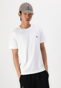 Maglietta bianca in cotone con colletto giro e maniche corte, con un piccolo logo navy sul petto. Abbinata a pantaloni corti scuri e un cappellino grigio.