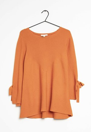 Haut à manches longues orange en maille côtelée avec encolure ronde et nœuds noués sur les manches, suspendu à un cintre noir sur un fond blanc.