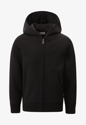 Persoon draagt een grijze sweater met print en joggingbroek, met een zwarte hoodie met rits, handen in de zakken, en witte sneakers tegen een effen achtergrond.
