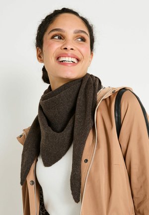 Mujer sonriendo con el cabello oscuro recogido en un moño, vistiendo una bufanda marrón, una chaqueta beige, una blusa blanca y un bolso negro sobre un fondo neutro.