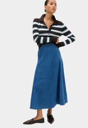 Mujer con camiseta de manga larga a rayas negras y blancas, falda vaquera azul hasta los tobillos y mocasines negros puntiagudos, de pie con los brazos cruzados.
