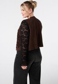 Blonde vrouw met een updo, gekleed in een bruine suède gilet, een zwart bloemen kant lange mouwen top, en hoge taille zwarte jeans, staande tegen een witte achtergrond.