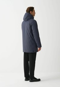 Muž v námorníckej kapucňovej parke, čiernych nohaviciach a čiernych topánkach, stojaci bokom na bielom pozadí.