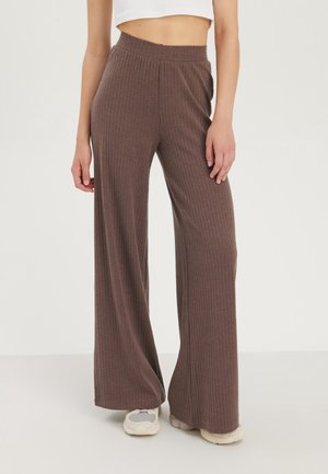 Donna che indossa pantaloni marroni a coste, a gamba larga e alta vita, e sneakers bianche, abbinati a un top corto bianco, in piedi su un pavimento bianco.